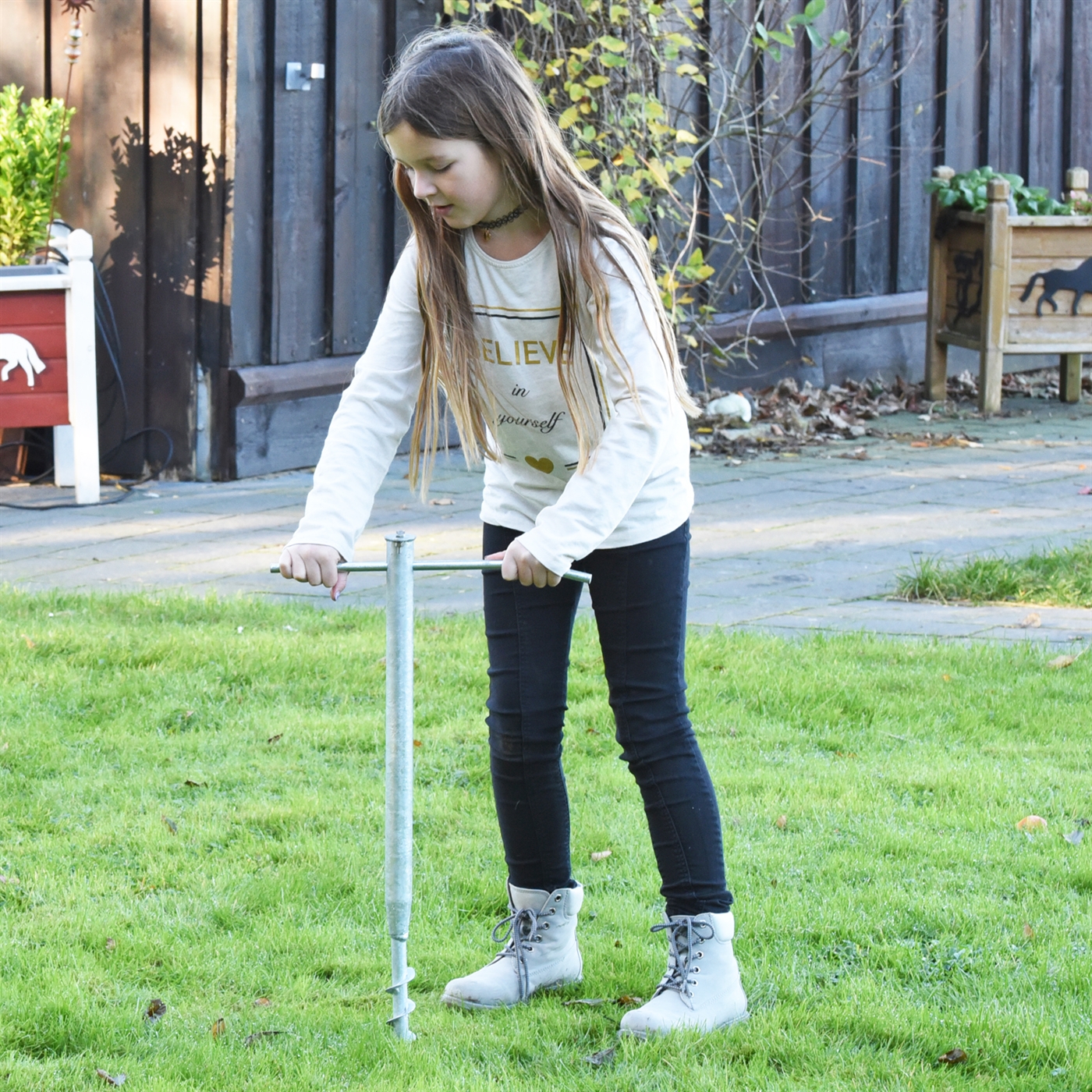 Ein Mädchen zeigt das Eindrehen des VOSS.farming Einschraub-Erdstab „Driller“ (80 cm) auf einer Grasfläche.