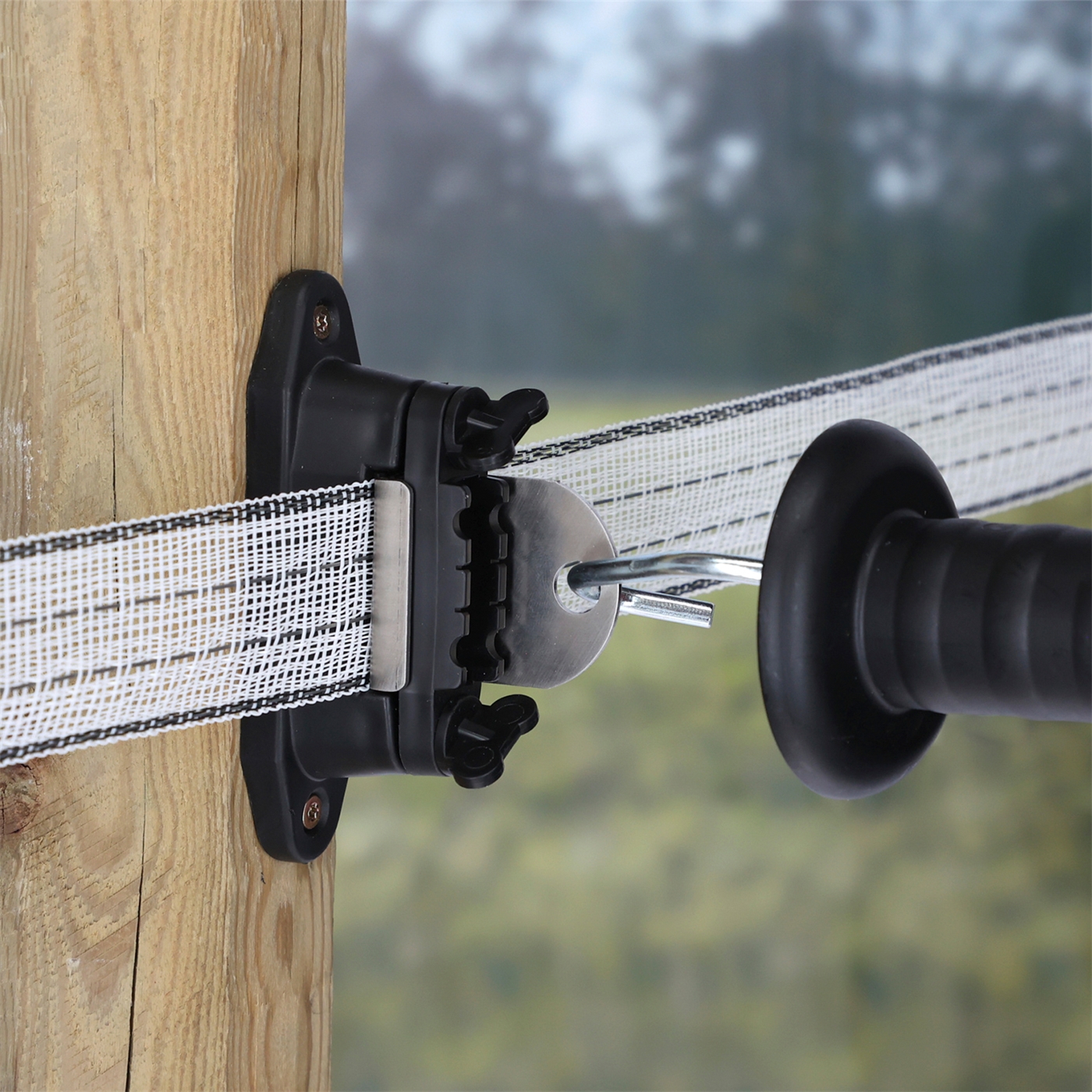Doppelklicken zum vergrößern Nahaufnahme eines Kerbl 3x Kombi-Torgriffisolator „Profi“ für T-Abtrennungen an Streckenpfahl und Ecken. Abbildung des an einem Holzpfahl angeschraubten Isolators, der ein Weidezaunband führt und in dessen Vorrichtung ein Torgriff eingehängt ist.