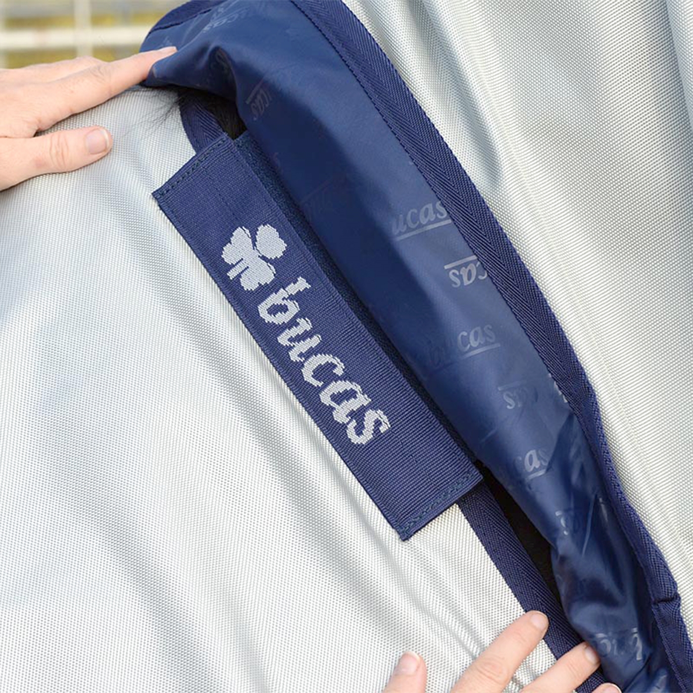 Doppelklicken zum vergrößern Nahaufnahme von Händen, die eine Bucas Power Turnout Medium 150 g, 125 cm Winterdecke für Pferde in Grau und Marineblau mit sichtbarem Markenlogo anpassen.