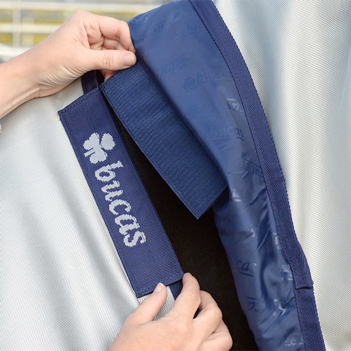 Doppelklicken zum vergrößern Hände, die einen blauen Klettverschluss mit dem Bucas-Logo an einer silbernen Bucas Power Turnout Medium 150 g, 125 cm Winterdecke für Pferde befestigen.