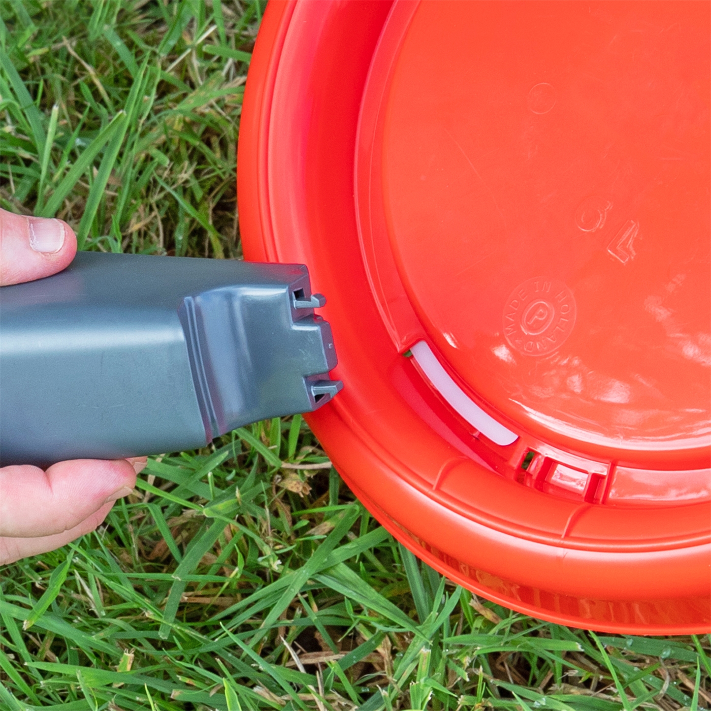 Doppelklicken zum vergrößern Eine Hand befestigt die „Geflügeltränke mit Bajonettverschluß, Inhalt 1,5 l“ – eine graue Geflügeltränke – am Rand eines umgedrehten orangefarbenen Plastikeimers auf einer Wiese.