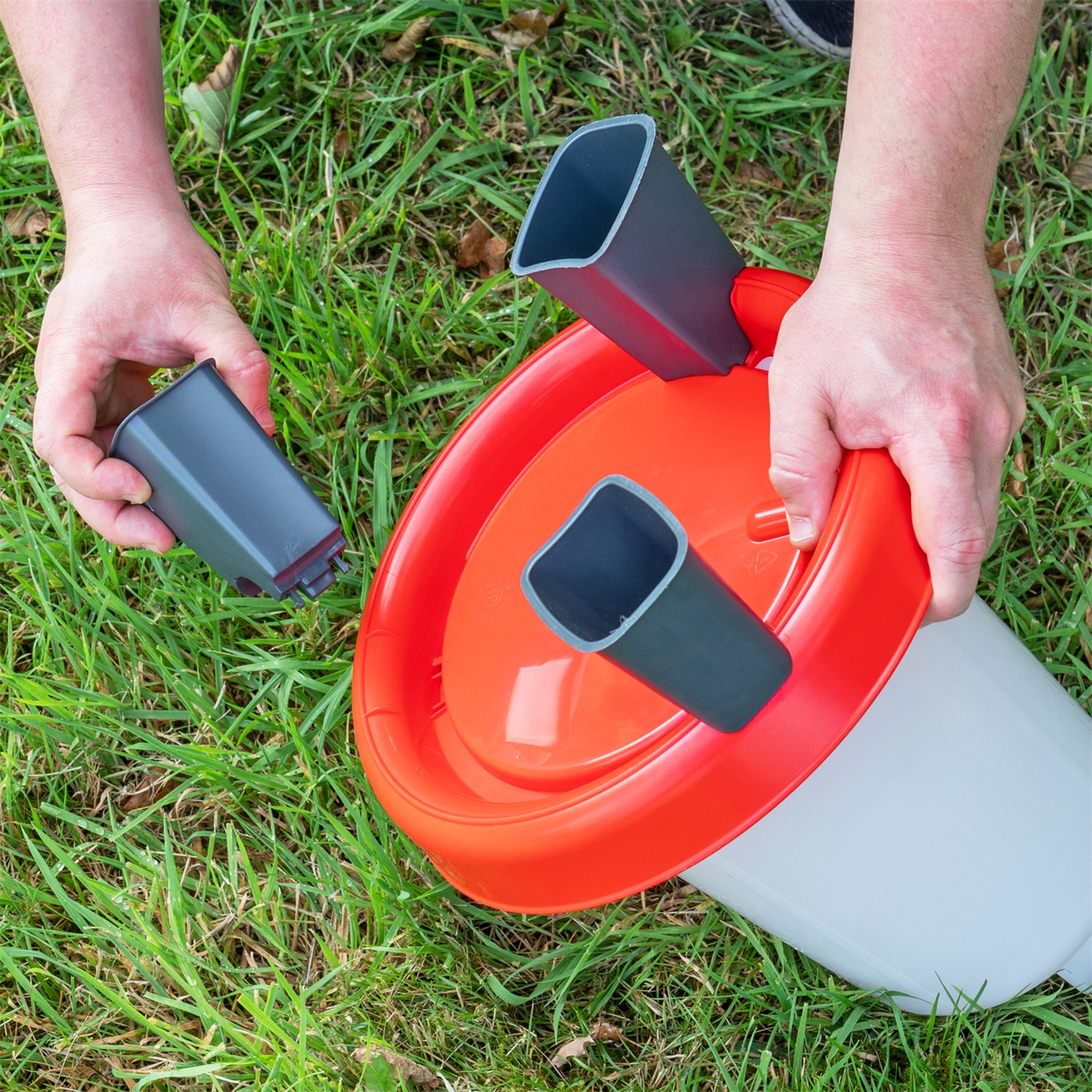 Doppelklicken zum vergrößern Hände montieren graue Plastikbeine an einer Geflügeltränke mit Bajonettverschluß, Inhalt 1,5 l – rot-weiße Geflügeltränke – auf grünem Gras im Freien.