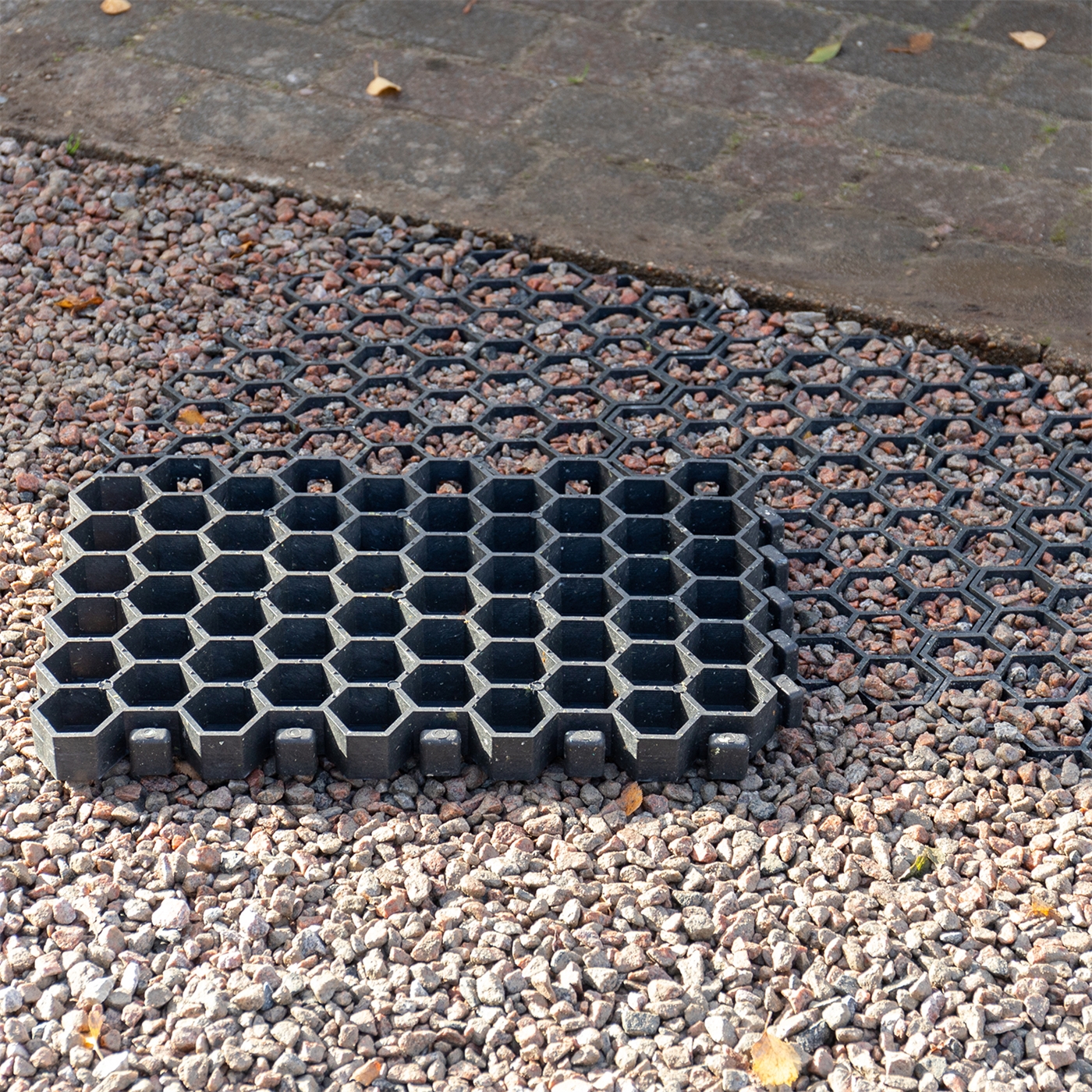 Doppelklicken zum vergrößern Paddockplatte Rasengitter (50x40x4cm), ein schwarzes sechseckiges Kunststoffgitter von VOSS.farming, ist teilweise auf Schotter gelegt, im Hintergrund befindet sich ein Ziegelpflaster.