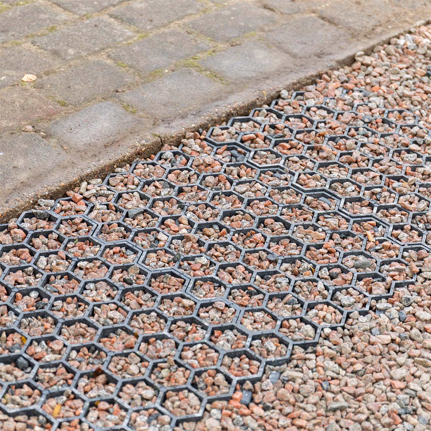 Doppelklicken zum vergrößern VOSS.farming Paddockplatte Rasengitter (50×40×4 cm) aus schwarzem Kunststoff mit sechseckigen Wabenzellen, die mit kleinen Steinen gefüllt sind. Die Platte liegt neben einem gepflasterten Gehweg.