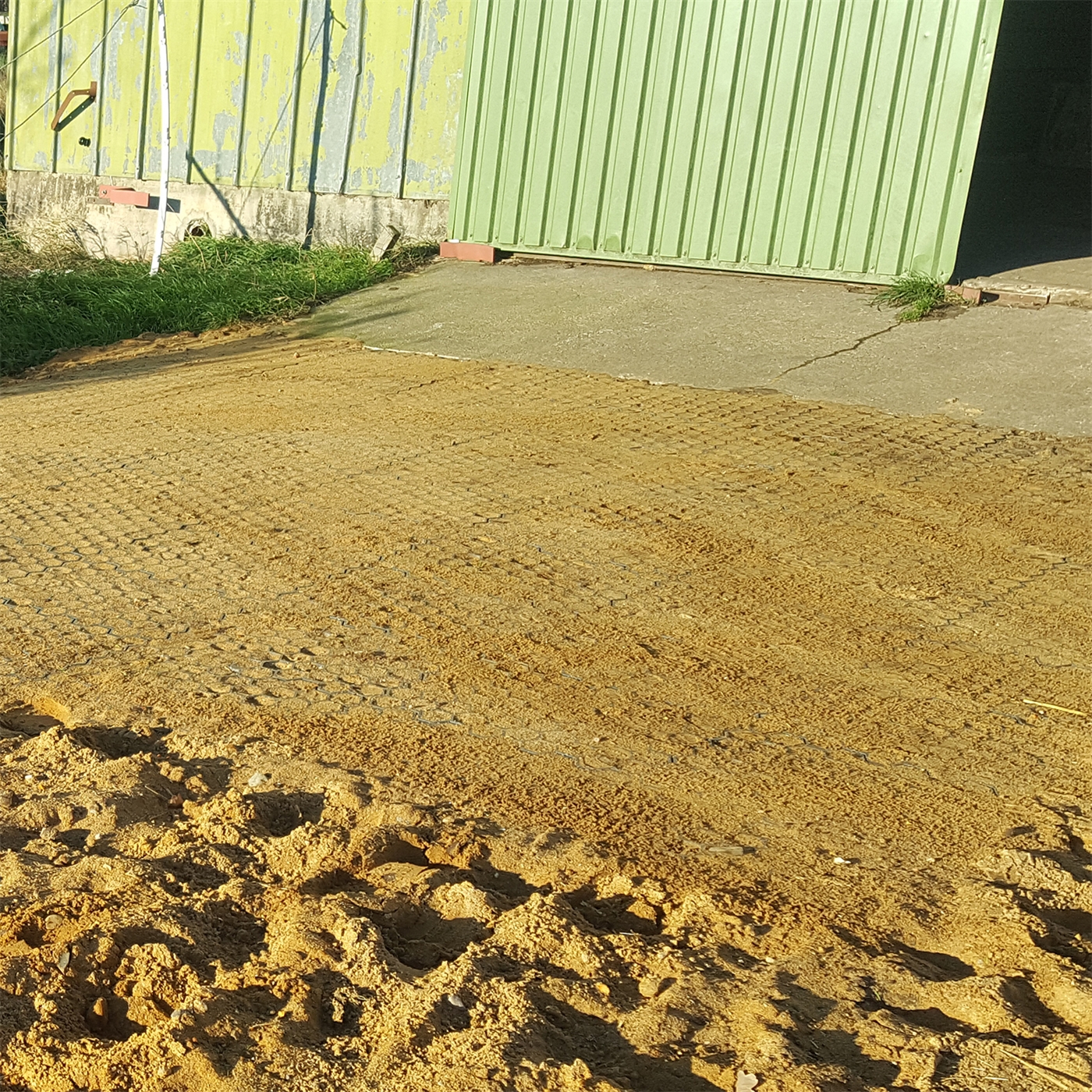 Doppelklicken zum vergrößern Paddockplatte Rasengitter (50x40x4cm) von VOSS.farming auf sandigem Boden mit Gittermuster neben einem grünen Metallschuppen und Betonweg im Sonnenlicht installiert.