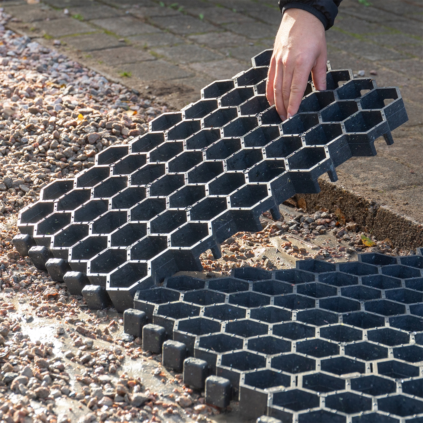 Doppelklicken zum vergrößern Eine Hand legt ein VOSS.farming Paddockplatte Rasengitter (50x40x4cm) auf Schotter neben Pflastersteinen.