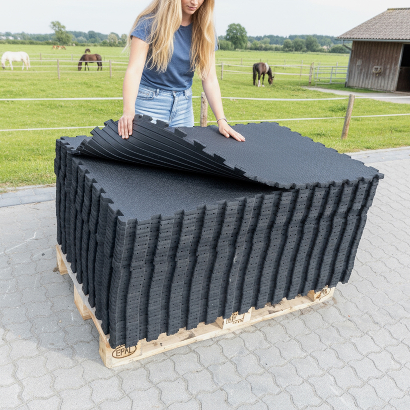 Eine Frau stapelt VOSS.farming Stallmatte Puzzlematte 120x90cm, 18mm Gummimatten auf einer Palette im Freien neben einem Feld mit Pferden.