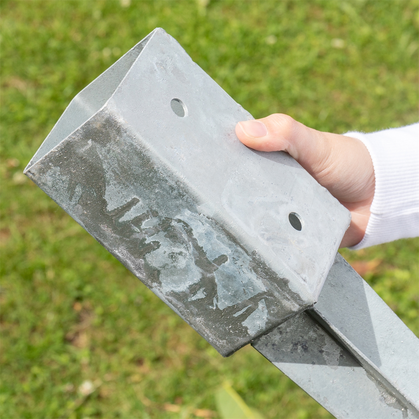 Eine Hand hält eine Bodeneinschlaghülse, Metall, 7 x 7 x 75cm, Halterung mit zwei Löchern, im Freien auf grünem Gras.