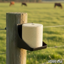 Ein S.I.N. Hellas VOSS.vital Leckstein - BiotinLine (5kg) Mineralleckstein mit Biotin & Vitamin E sitzt auf einem Zaunpfahl auf einer Wiese, während im Hintergrund Kühe grasen.