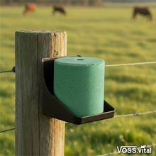 Ein VOSS.vital Leckstein - KräuterLine Mineral-Leckstein mit Kräutern 5kg von S.I.N. Hellas an einem Pfahlhalter in der Nähe eines Stacheldrahtzauns auf einer Weide mit grasenden Kühen.