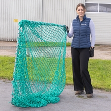 Eine Frau steht neben einem VOSS.farming Rahmen für Futtersparnetz – für quadratische Futterraufen, 1,68 m x 1,68 m, verzinkt, einem großen, quadratischen Metallrahmen, der mit einem grünen Maschennetz bespannt ist.