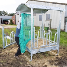 Eine Person, die ein grünes Heusparnetz in einen VOSS.farming Rahmen für Futtersparnetz – für quadratische Futterraufen, 1,68 m x 1,68 m, verzinkt einlegt, bei dem es sich um einen quadratischen, verzinkten Metall-Futtertrog mit Dach handelt.