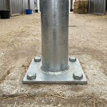 Nahaufnahme des Growi® Anbindepfostens 130cm Rundrohr (Ø 102mm) mit Bodenplatte, sicher verschraubt auf einem Stallboden, im Hintergrund Ställe und Heu. Ideal für Pflege- oder Waschplätze.