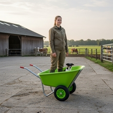 Eine Frau steht in einer LandXpert Schubkarre – 250 Liter, grün, mit Doppelbereifung –, in der auch ein kleiner Hund sitzt, auf einem Bauernhof.