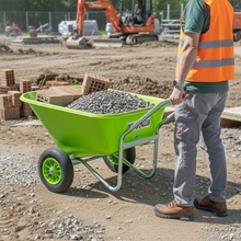 Ein Arbeiter in einer orangefarbenen Weste steht neben einer LandXpert-Schubkarre – 250 Liter, grün, mit Doppelbereifung, Kunststoffwanne und Luftreifen, gefüllt mit Kies und Ziegeln – auf einer Baustelle.