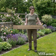 Eine Person steht in einem Garten und hält einen VOSS.garden Steckzaun 75x18cm, eine Holzeinfassung mit Metallpfosten für Rasenkanten; Blumen und Grünzeug umgeben sie.