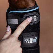 Eine Person befestigt die Waldhausen Health + Care Gamasche von Waldhausen, eine schwarze Beinorthese mit Kühl-, Wärme- und Vibrationsfunktion, mit Klettbändern auf einem Ziegelsteinuntergrund am Bein eines Pferdes.