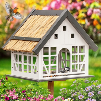 Das VOSS.garden „Lindau“ Vogelhaus im Fachwerkstil verfügt über ein weißes Design mit Strohdach, ist von Blumen umgeben und zeigt zwei Vögel – einen fliegenden und einen sitzenden.