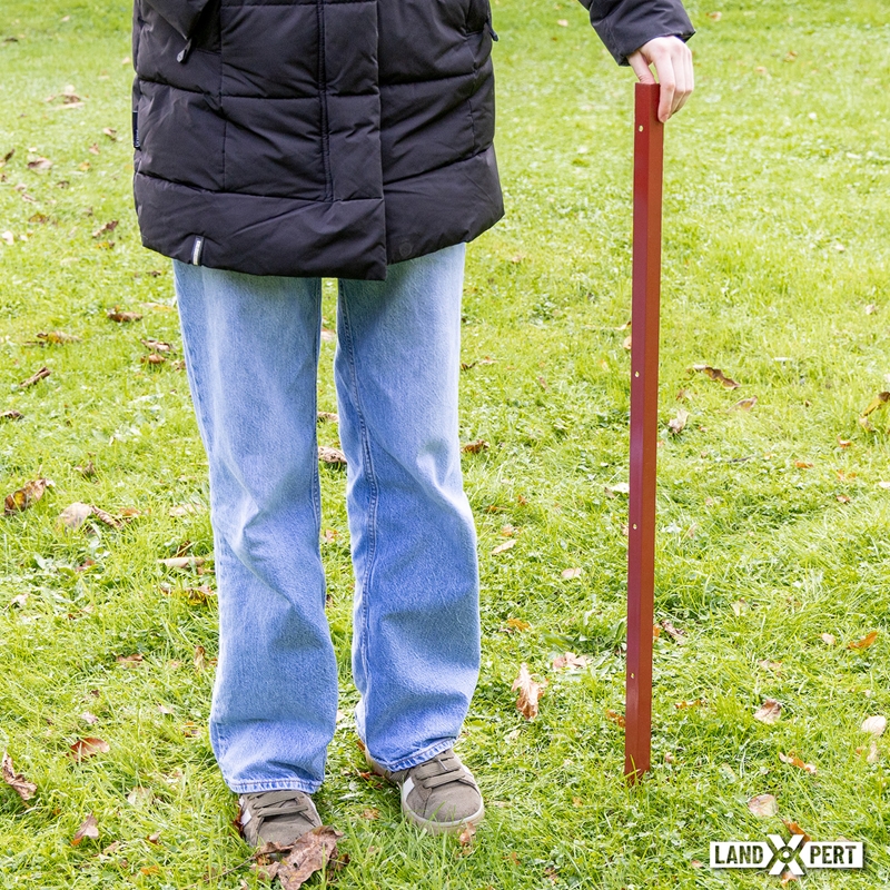 Großansicht der Bildergalerie Eine Person hält einen hohen, roten, L-förmigen LandXpert Winkelstahlpfahl (115 cm, 2 mm) aus Metall senkrecht auf einem Rasen.